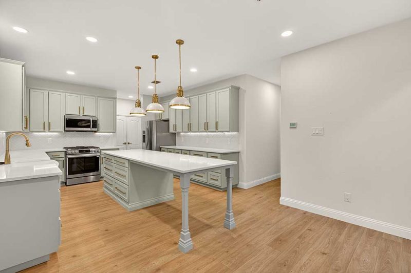 Modern kitchen with sage cabinets, brass fixtures, white island, wood floors, and stainless appliances.