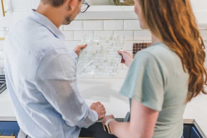 Two people review a glass tile sample in a bright kitchen with a white subway tile backsplash.